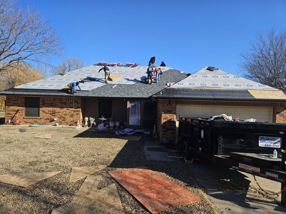 Roofers replacing shingles on a brick house under a blue sky, with a trailer parked in the driveway.