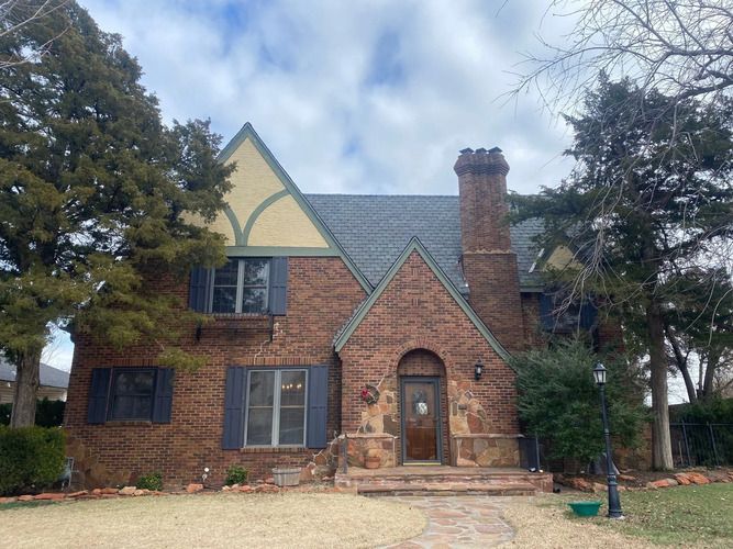 Brick house with dark blue shutters, chimney, and gray roof against a cloudy sky.