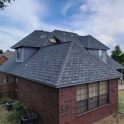 Two-story brick house with dark gray shingle roof under a partly cloudy sky.
