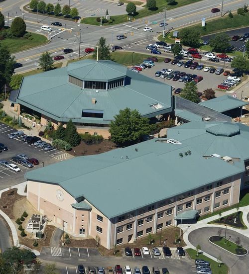 Aerial view of two-story light tan buildings with teal roofs, connected by a walkway. Parking lots and roads surround.