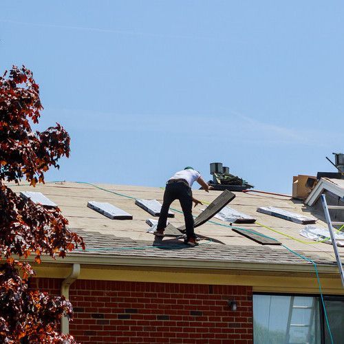 Roofer working on a roof, with scattered shingles. Blue sky and brick house visible.