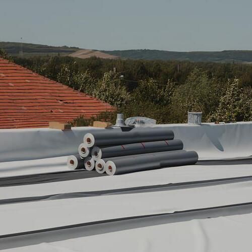 Rolls of roofing material on a flat roof with a red-tiled roof and trees in the background.