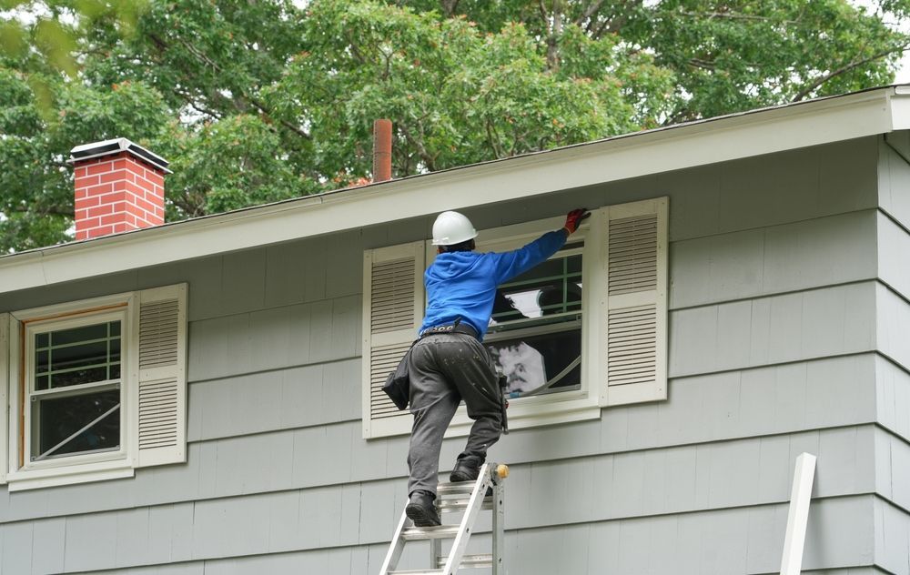 Person on ladder painting window trim on a gray house with white shutters.