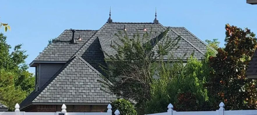 Grey-shingled roof of a house with decorative finials. Trees and a white fence are in the foreground.