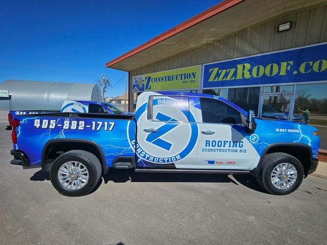 Blue and white truck with roofing company logo parked in front of building. 