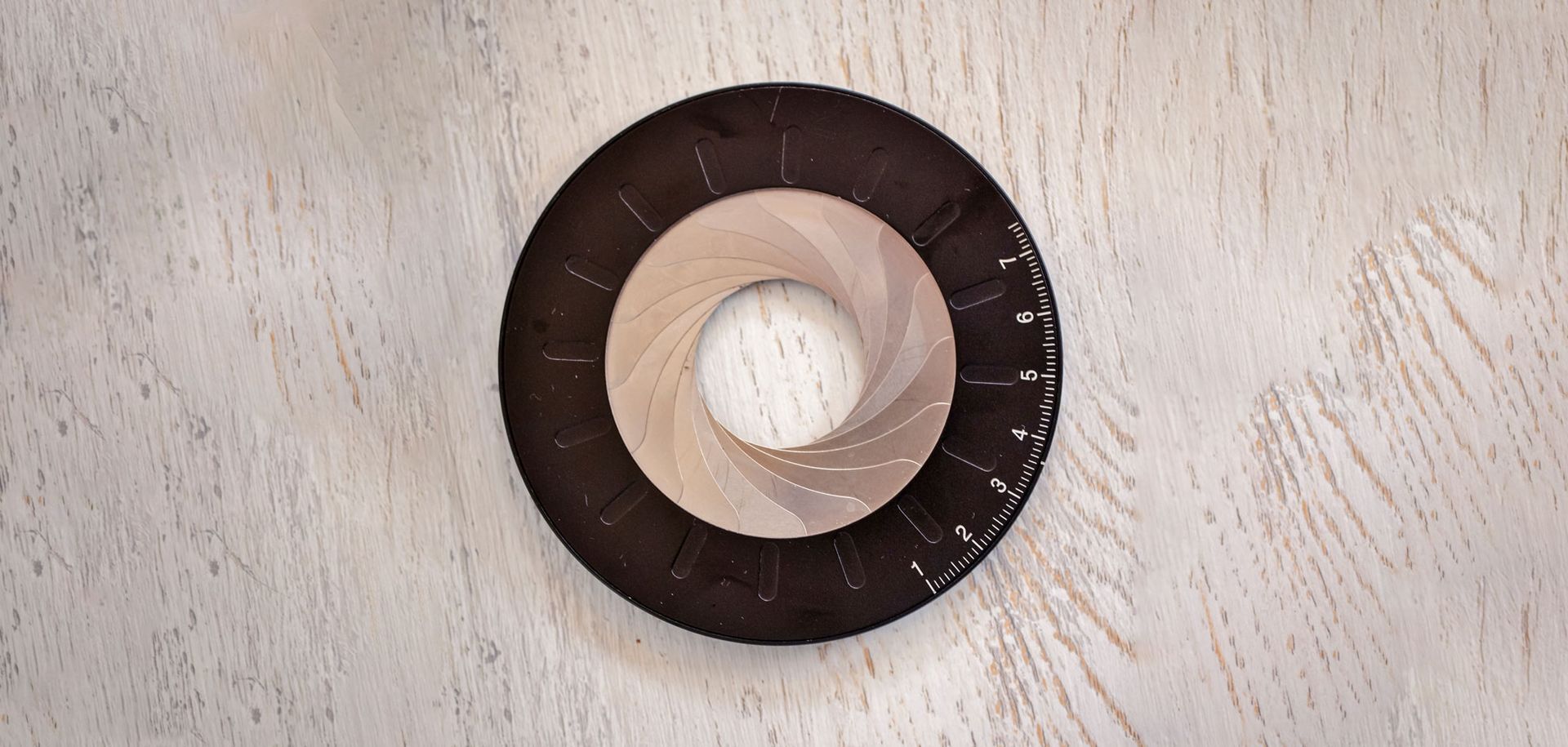 A mechanical aperture diaphragm with tan metal leaves, set within a black ring on a light-colored wood surface.