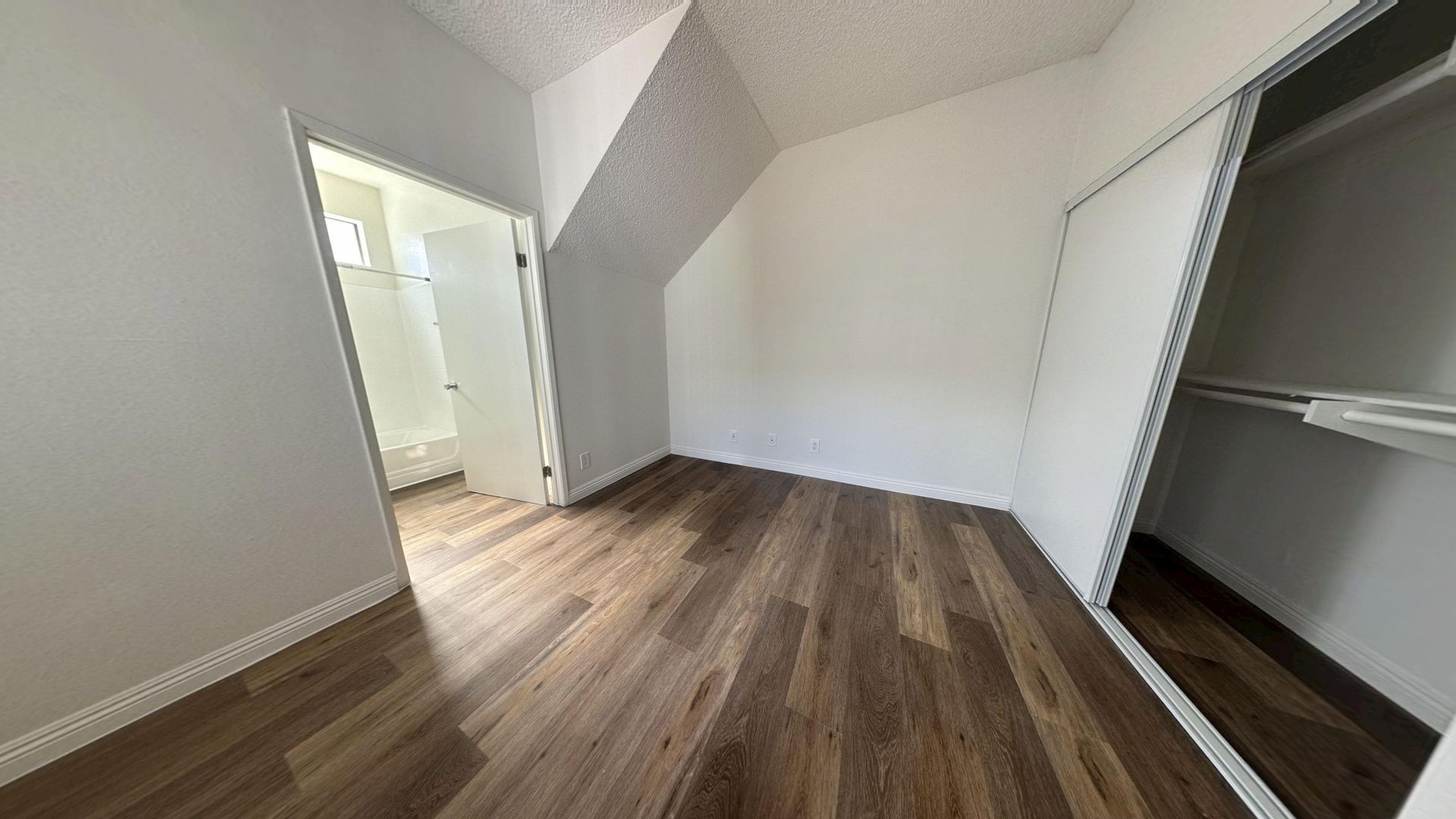 Empty bedroom with wood-look flooring, sloped ceiling, closet, and open doorway to bathroom.