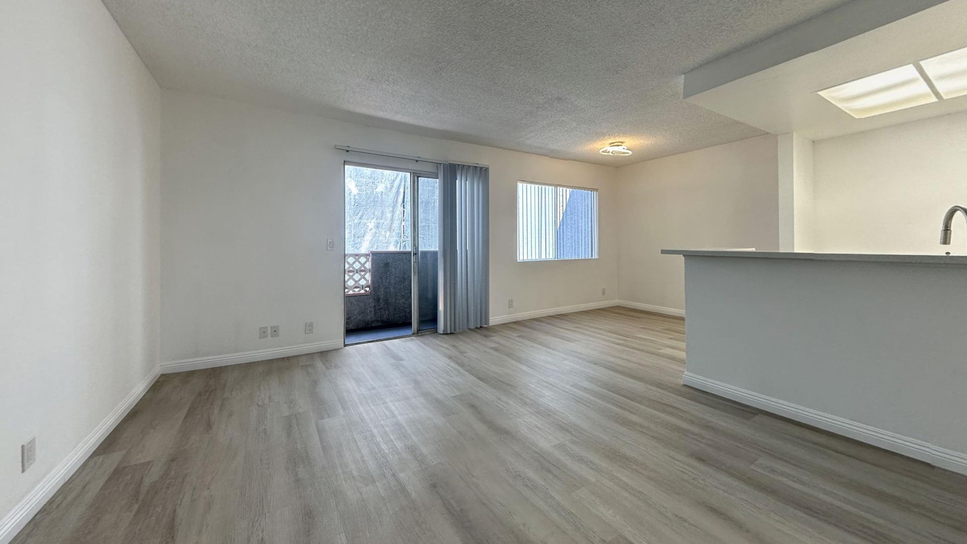 Empty apartment interior with wood-look flooring, balcony door, and kitchen island. White walls.