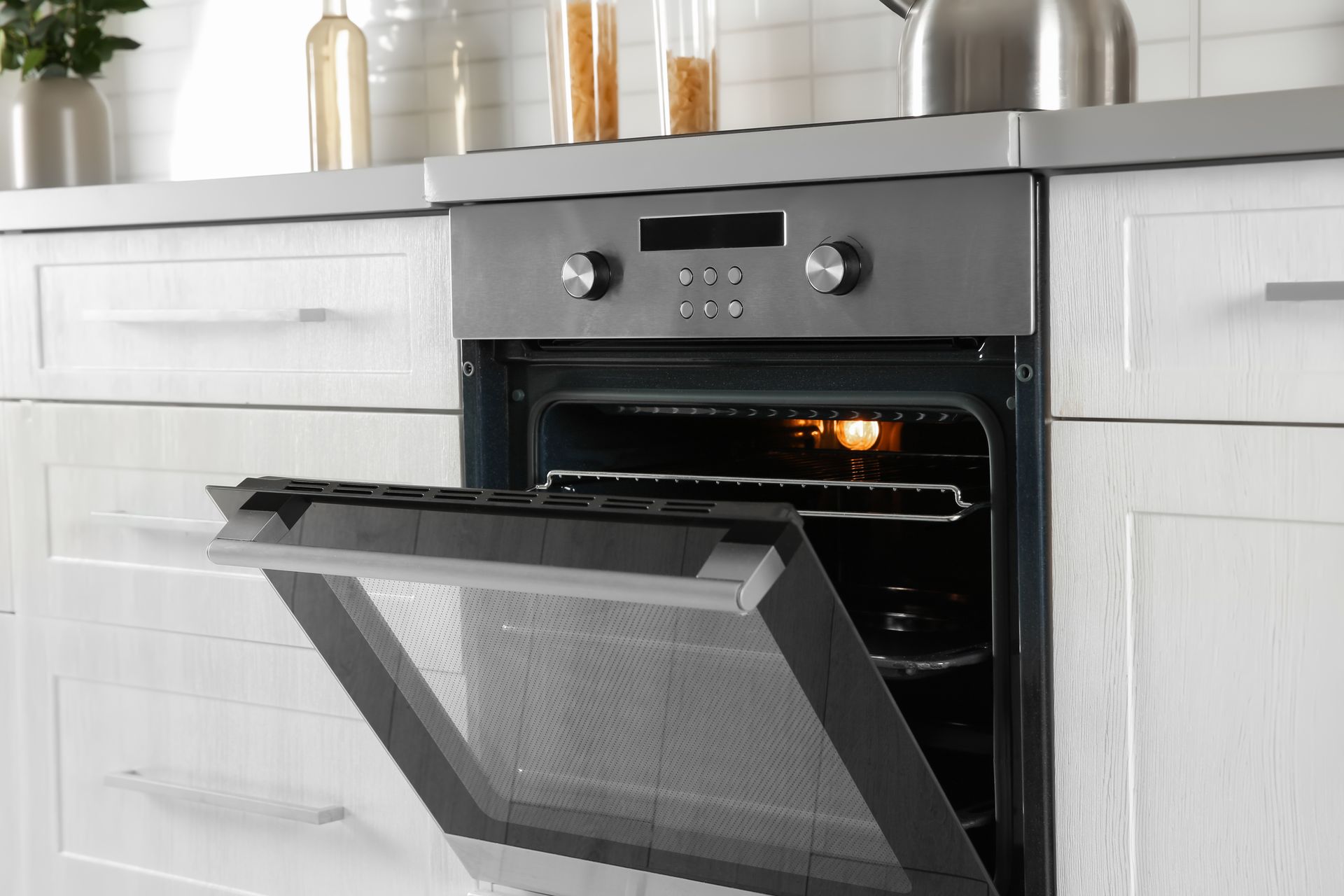 Oven with open door, in a white kitchen setting. Interior light is on, stainless steel face.