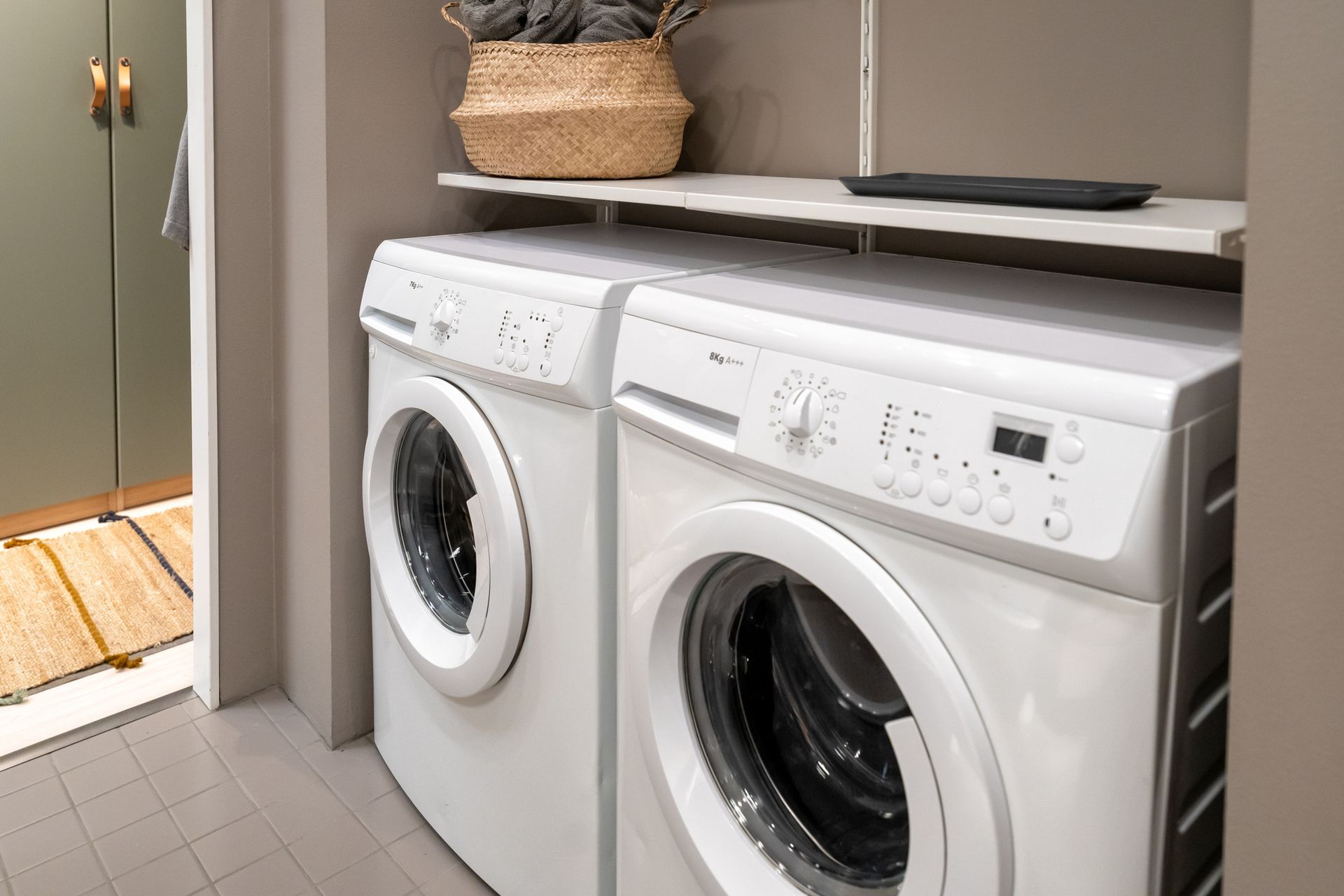 White washer and dryer units in a laundry room, with a shelf above holding a basket and a tray.