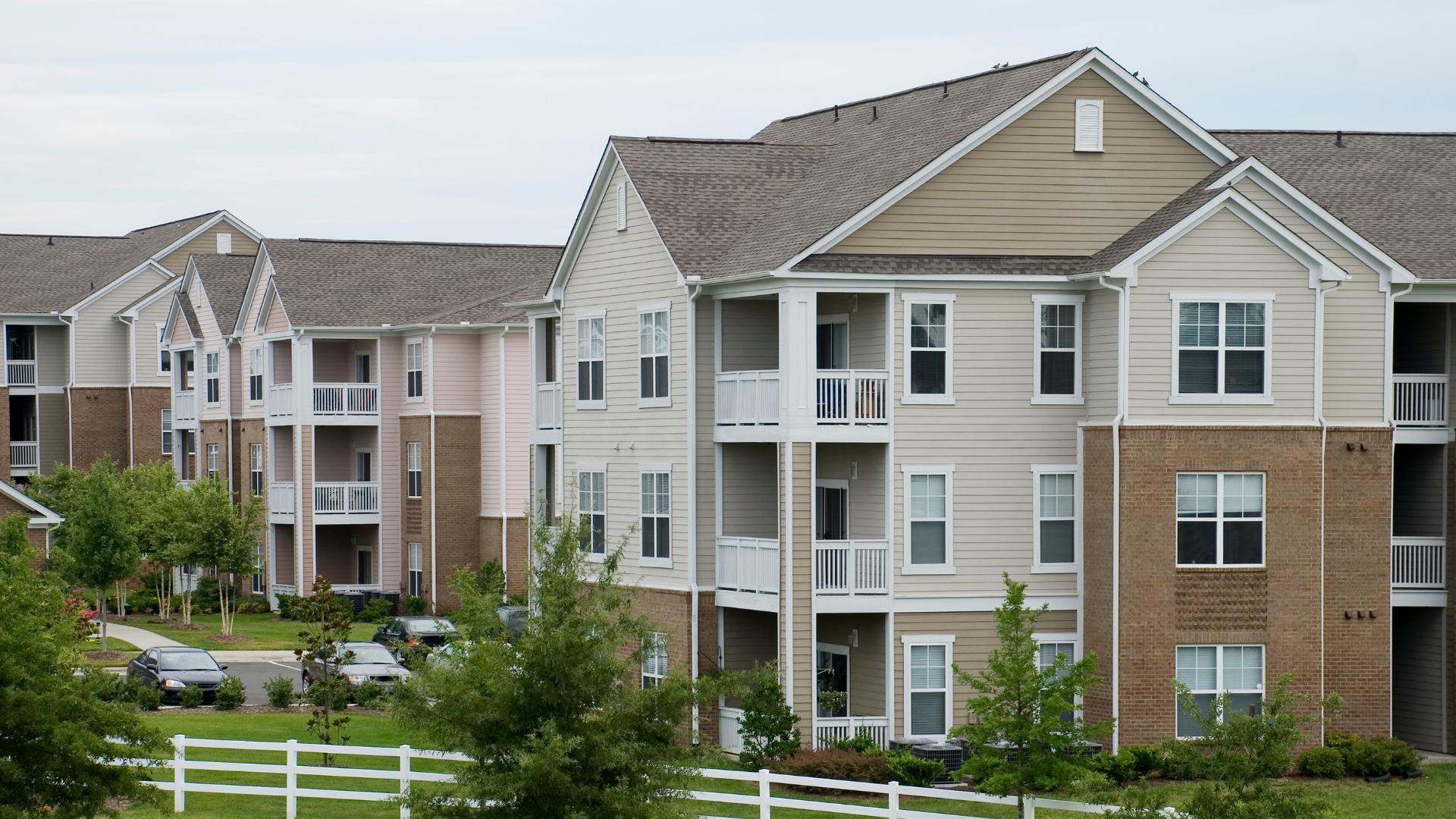 Apartment buildings with balconies, beige and brick facade, set behind a white fence and greenery.