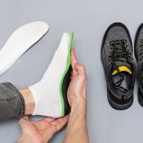 Una persona tiene in mano una soletta bianca e verde vicino a un paio di scarpe da ginnastica scure su uno sfondo grigio chiaro.