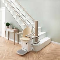 In una stanza luminosa, un montascale color crema è installato su una rotaia curva alla base di una scala in legno bianco.