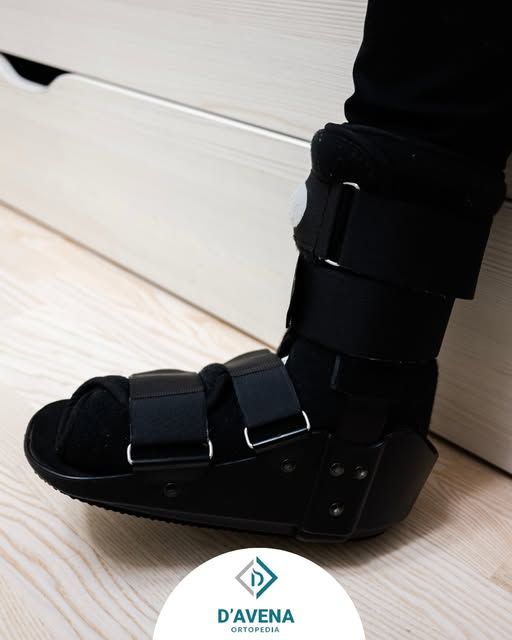 Una persona che indossa uno stivale ortopedico nero sopra un calzino nero, appoggiata a una superficie di legno chiaro.