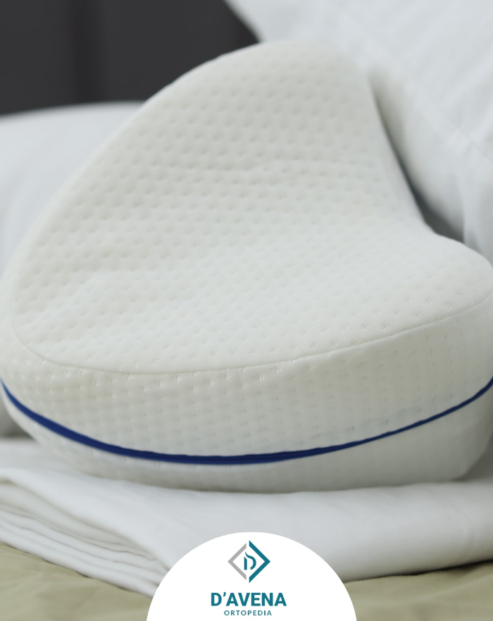 Un cuscino ortopedico per le gambe, bianco e a forma di cuore, con bordino blu, poggiato su lenzuola bianche con il logo D'Avena sottostante.