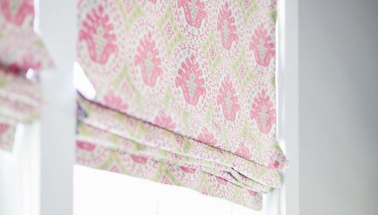 A close up of a pink and white roman shade on a window.