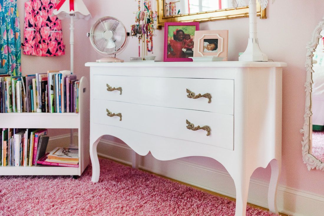 A white dresser is sitting in a pink room next to a bookshelf.