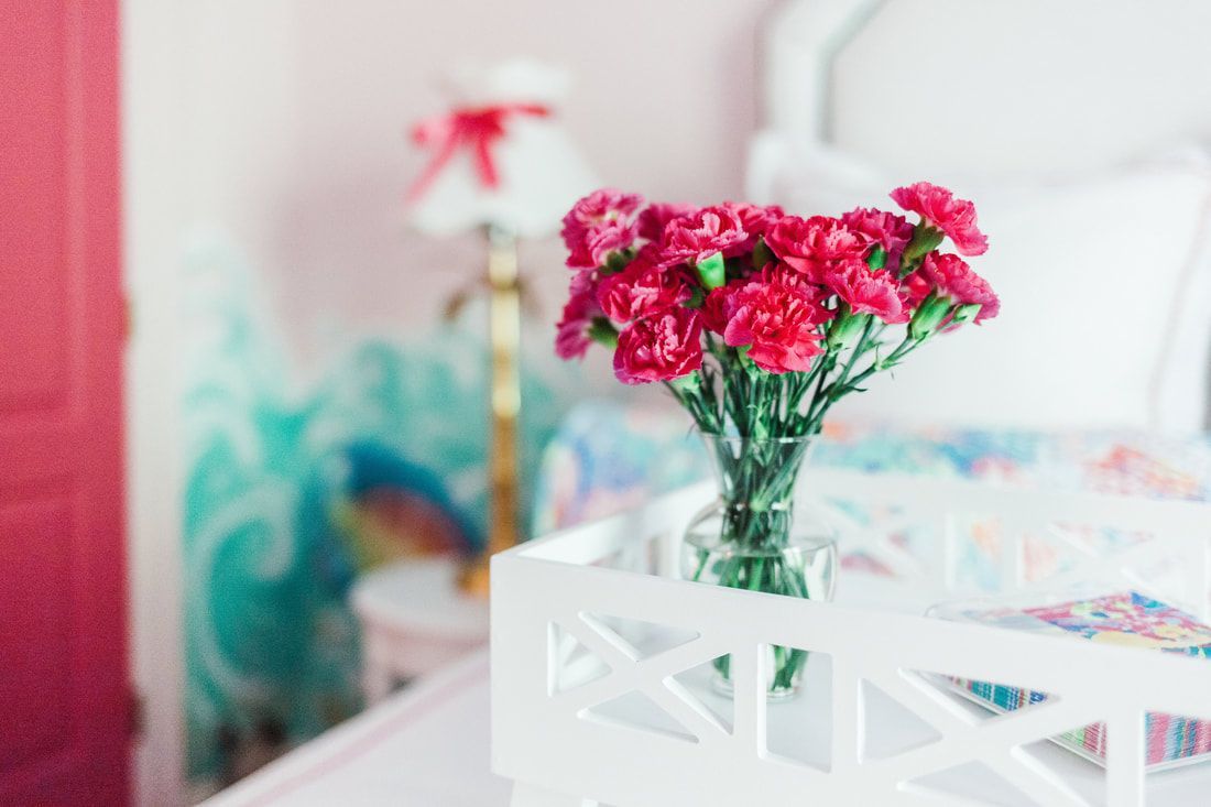 A vase of red carnations is sitting on a white table next to a bed.