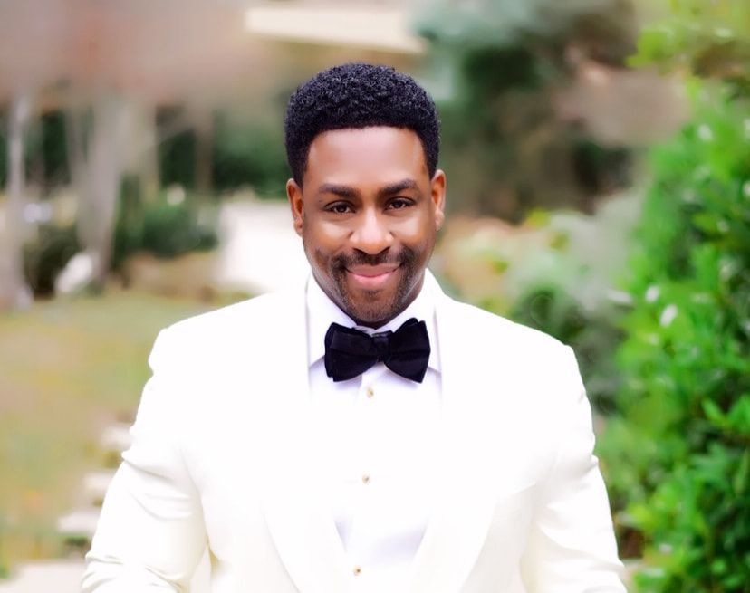 A man in a white suit and black bow tie is smiling.