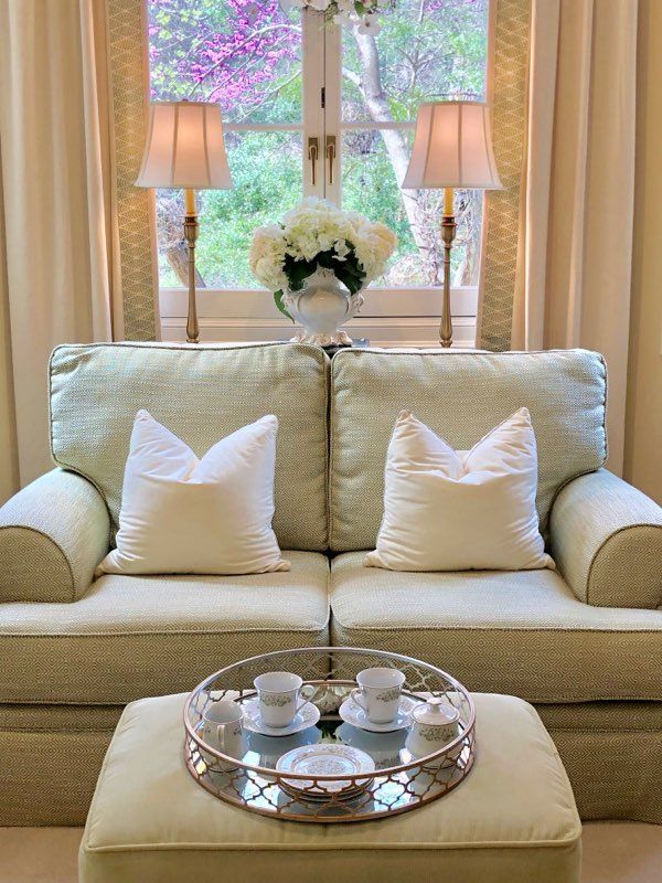 A living room with a couch , ottoman , lamps and a tray with cups and saucers on it.