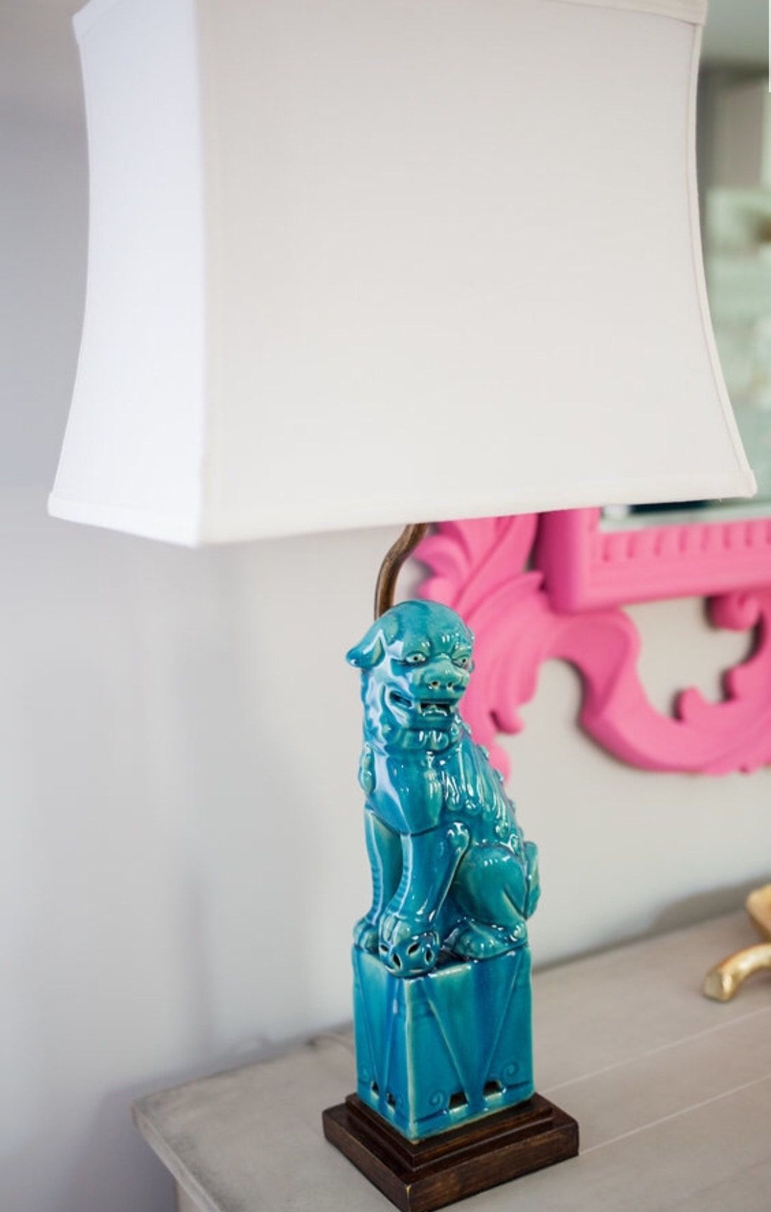 A blue lamp with a white shade is sitting on a table next to a pink mirror.