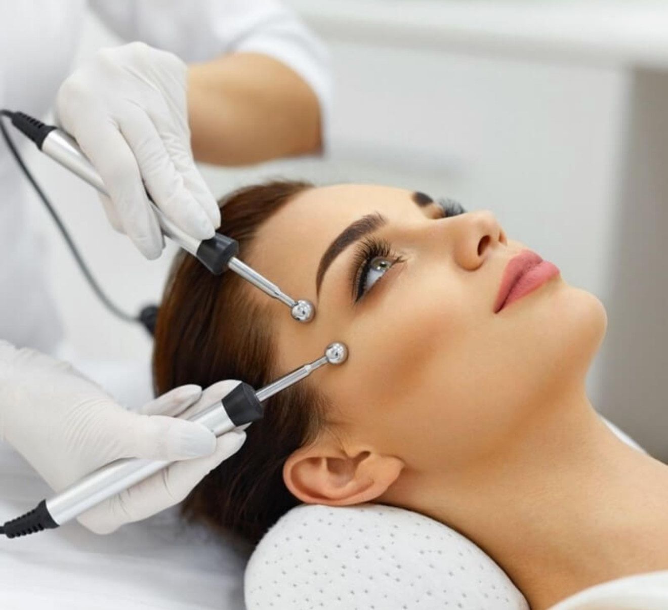 Woman receiving facial treatment with electrodes. Hands in gloves hold the devices over her face.