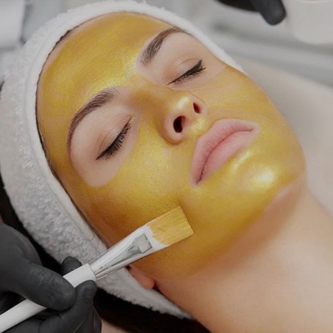 Woman with gold face mask, receiving a spa treatment, being painted on by gloved hands with a brush.