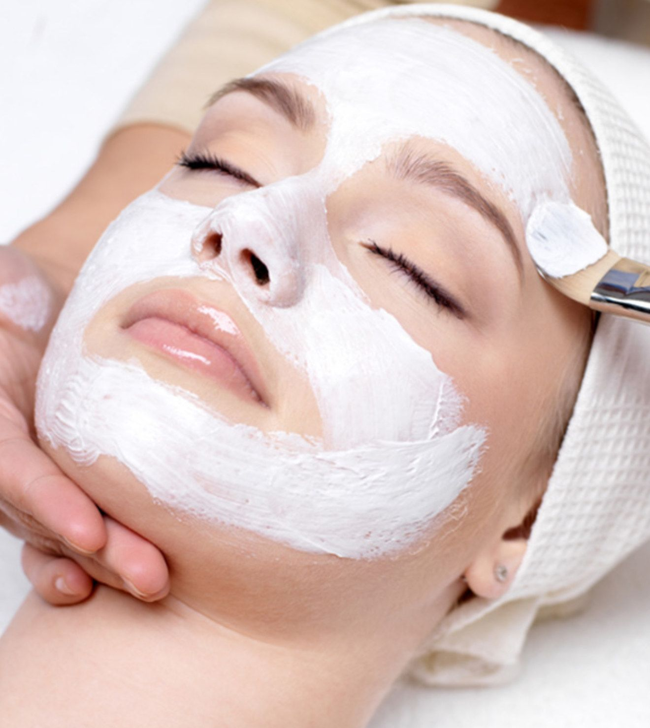 Woman receiving a facial mask with a brush, eyes closed, spa setting.