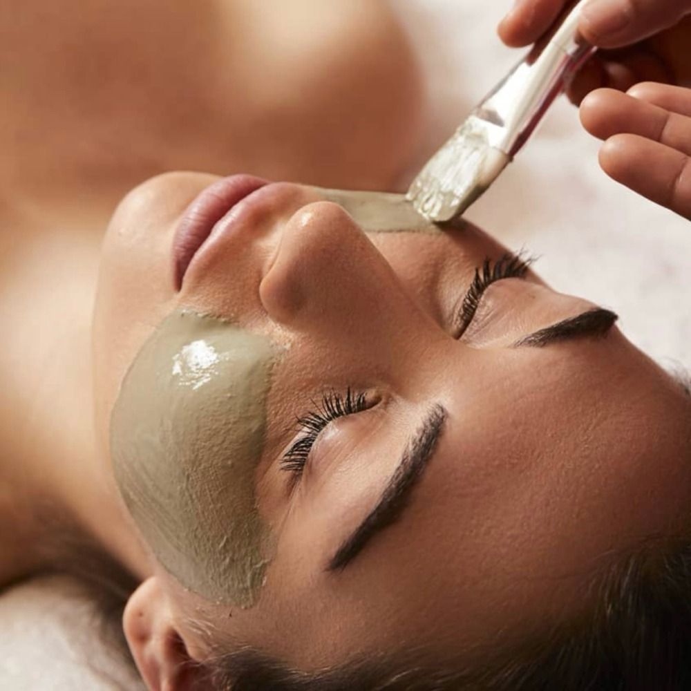 Woman receiving facial treatment; esthetician applying a clay mask with a brush.