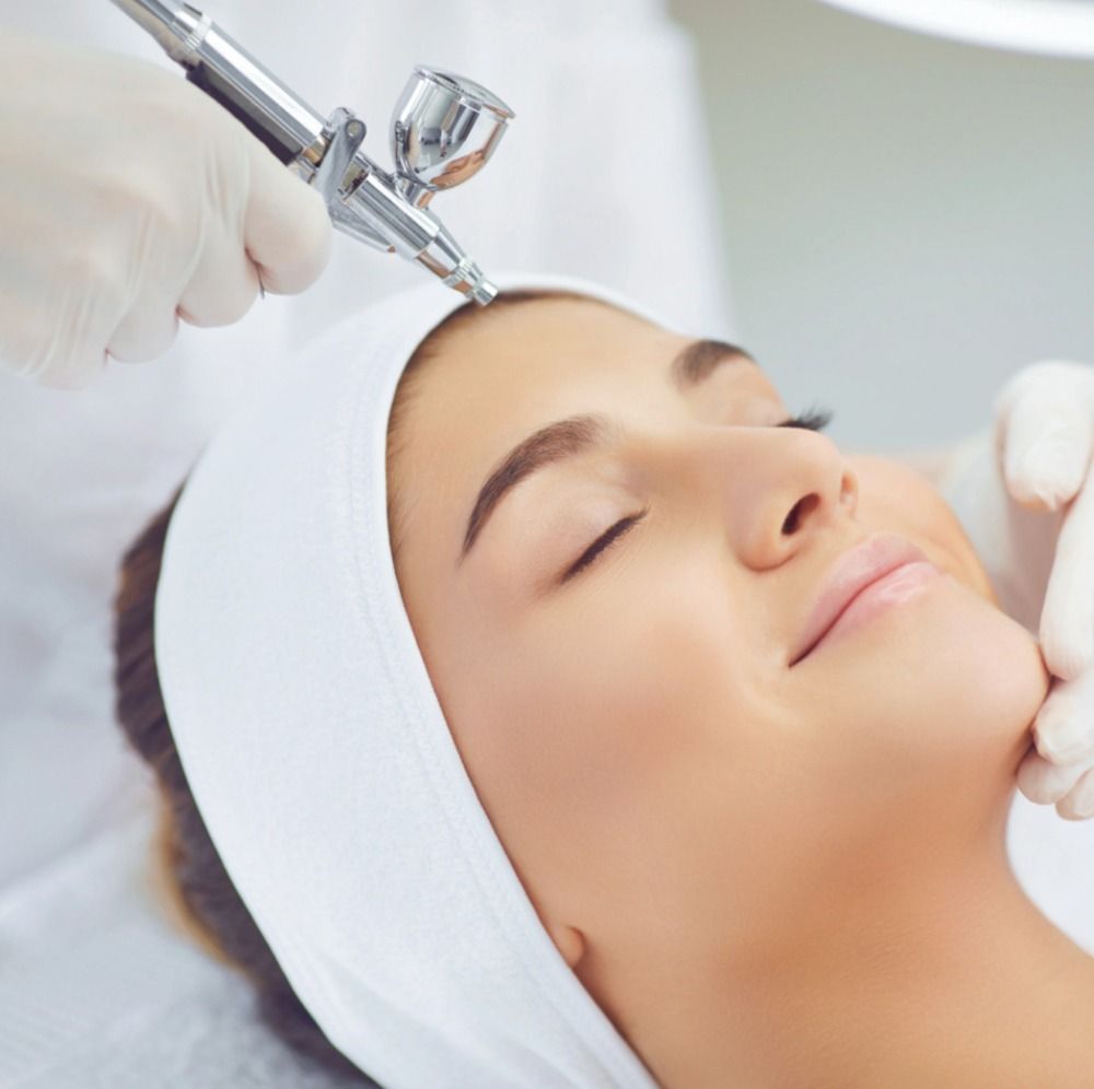Woman receiving oxygen facial treatment in spa, white headband, gloved hands, airbrush tool.