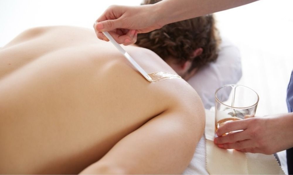 Person receiving back wax, a technician applying wax with a spatula. A glass of liquid sits nearby.