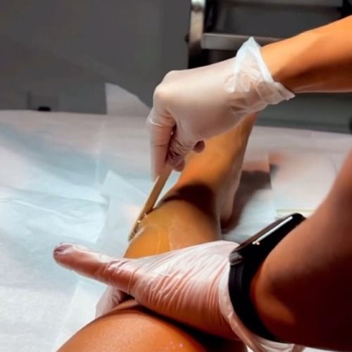Person's leg being waxed by gloved hands with wooden stick, in a salon setting.