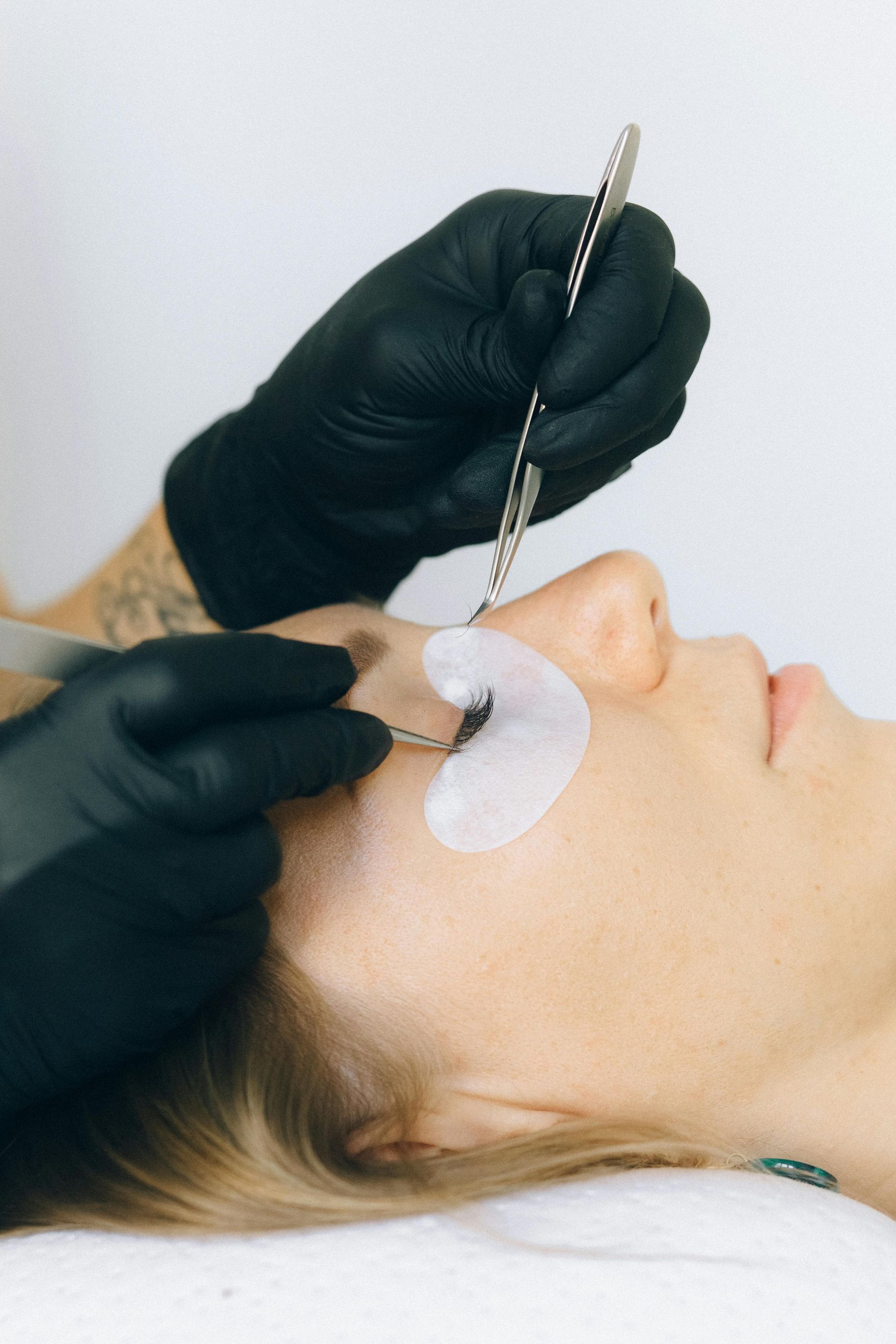 Eyelash extension application: technician's gloved hands using tools on a woman's eye with protective pads.