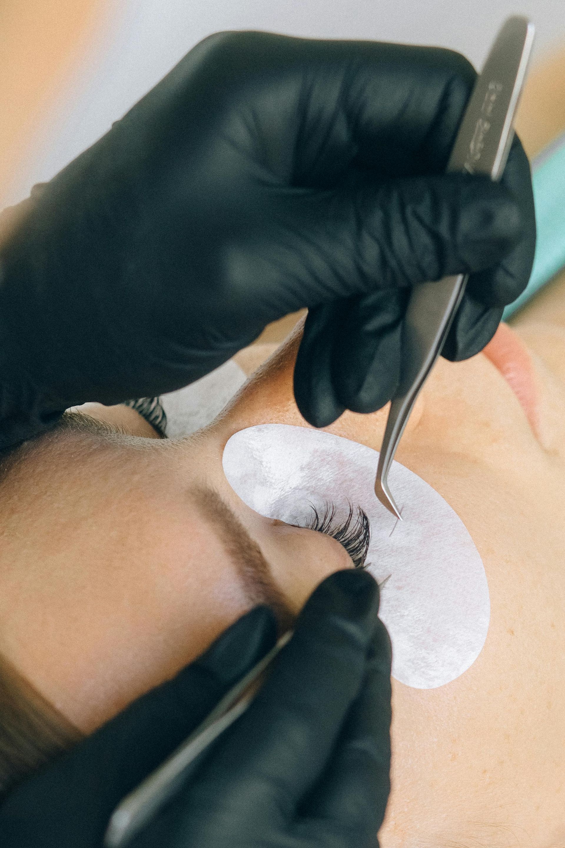 A person having eyelash extensions applied; black gloved hands hold tweezers near eye.