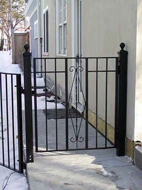 A wrought iron gate is leading to a snowy sidewalk in front of a house.