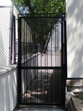 A black wrought iron gate leading to a walkway next to a chain link fence.