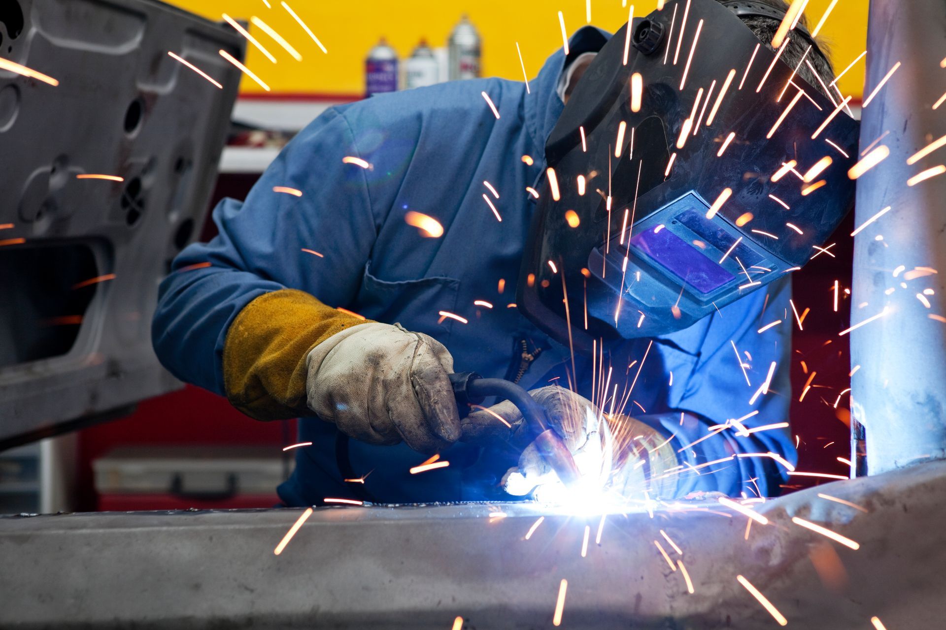 Welder in blue coveralls, helmet, and gloves welding metal with bright sparks in a shop.