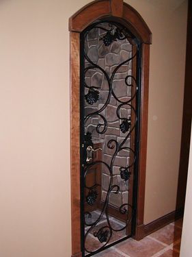 A wrought iron door with grapes on it is in a hallway next to a stone wall.