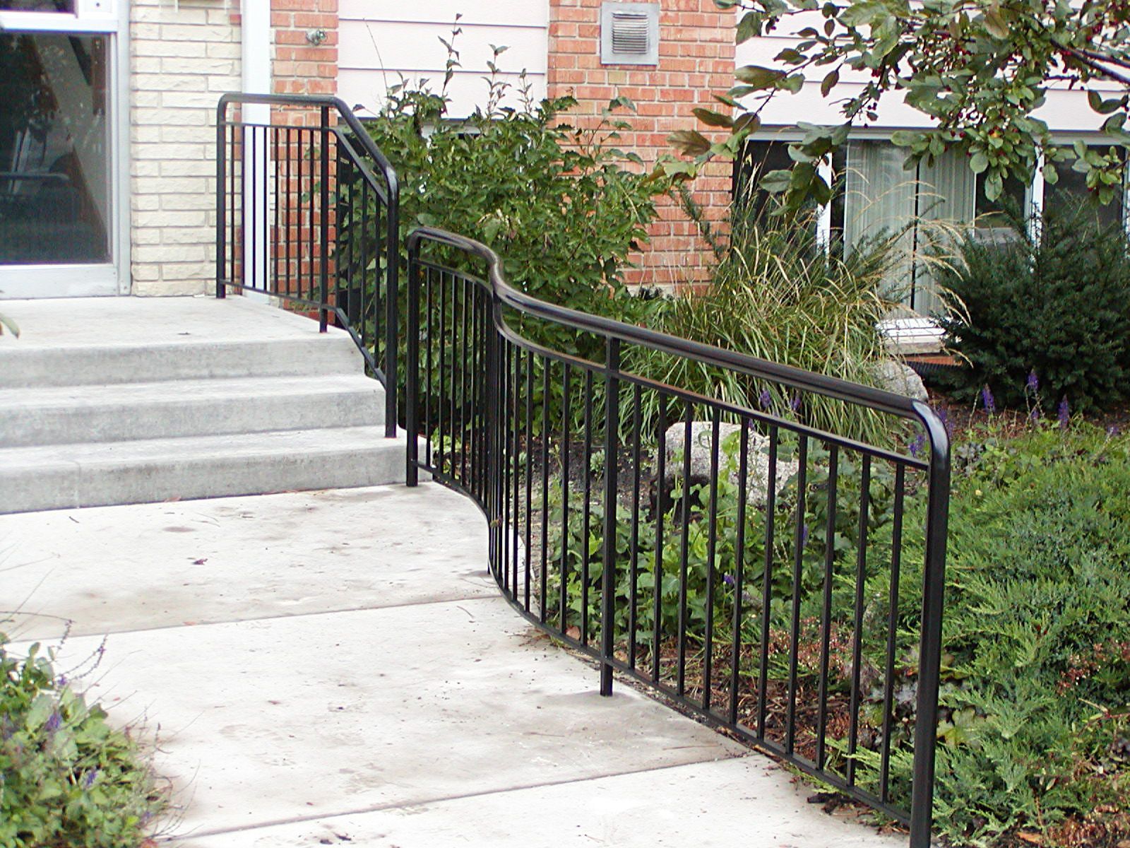 Black metal handrail alongside steps and a curved sidewalk in front of a building with greenery.