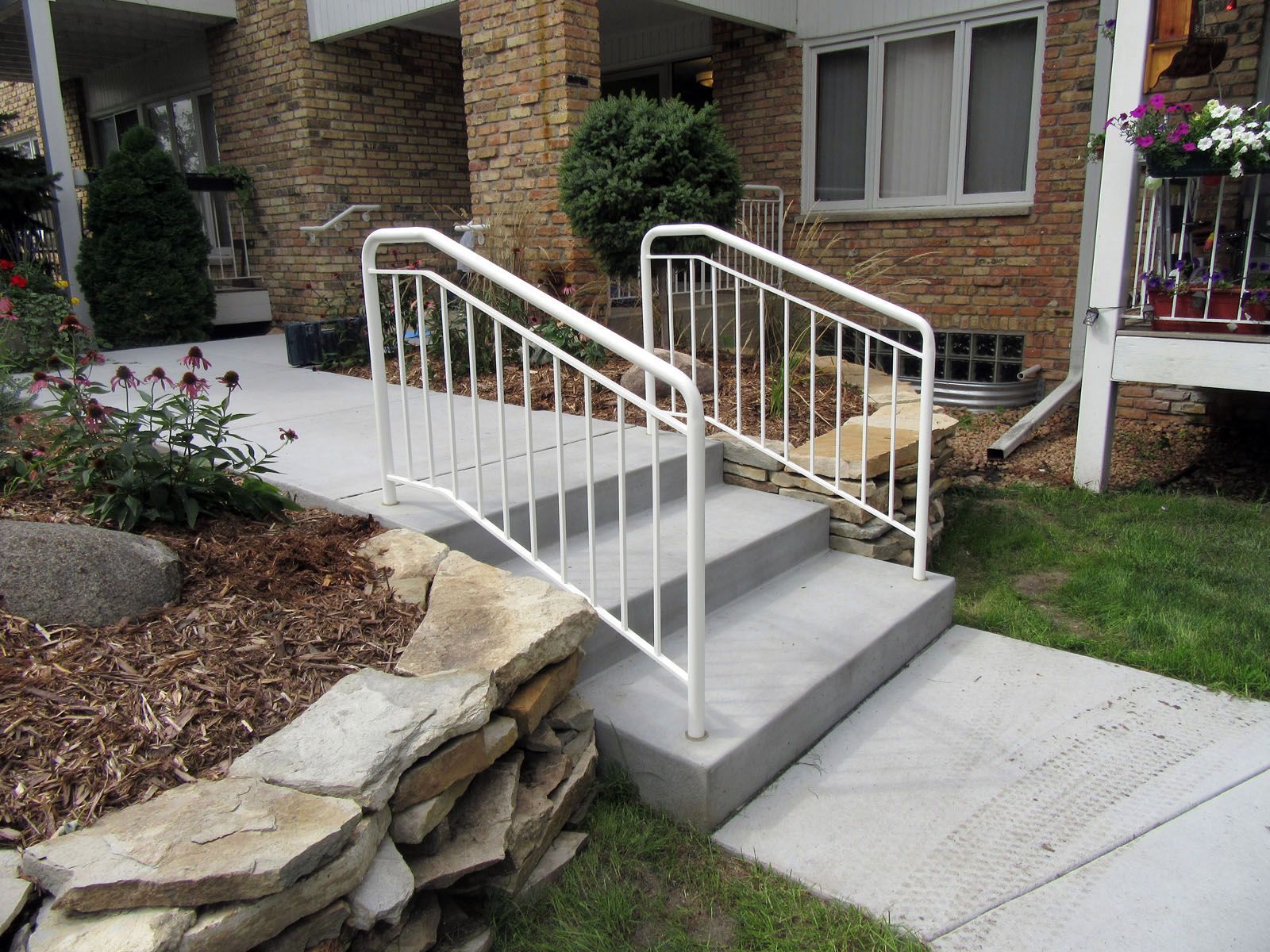 White handrails on concrete steps leading to a building entrance. A stone wall and landscaping are in the foreground.