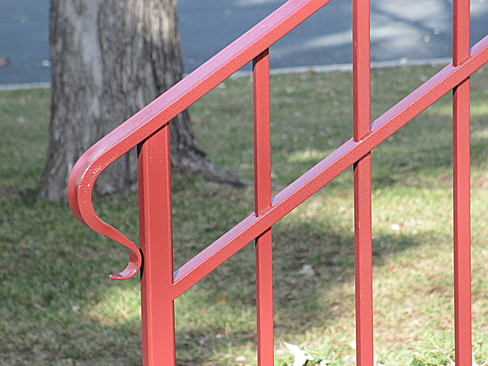 Red metal handrail with angled bars, against a tree and grass.