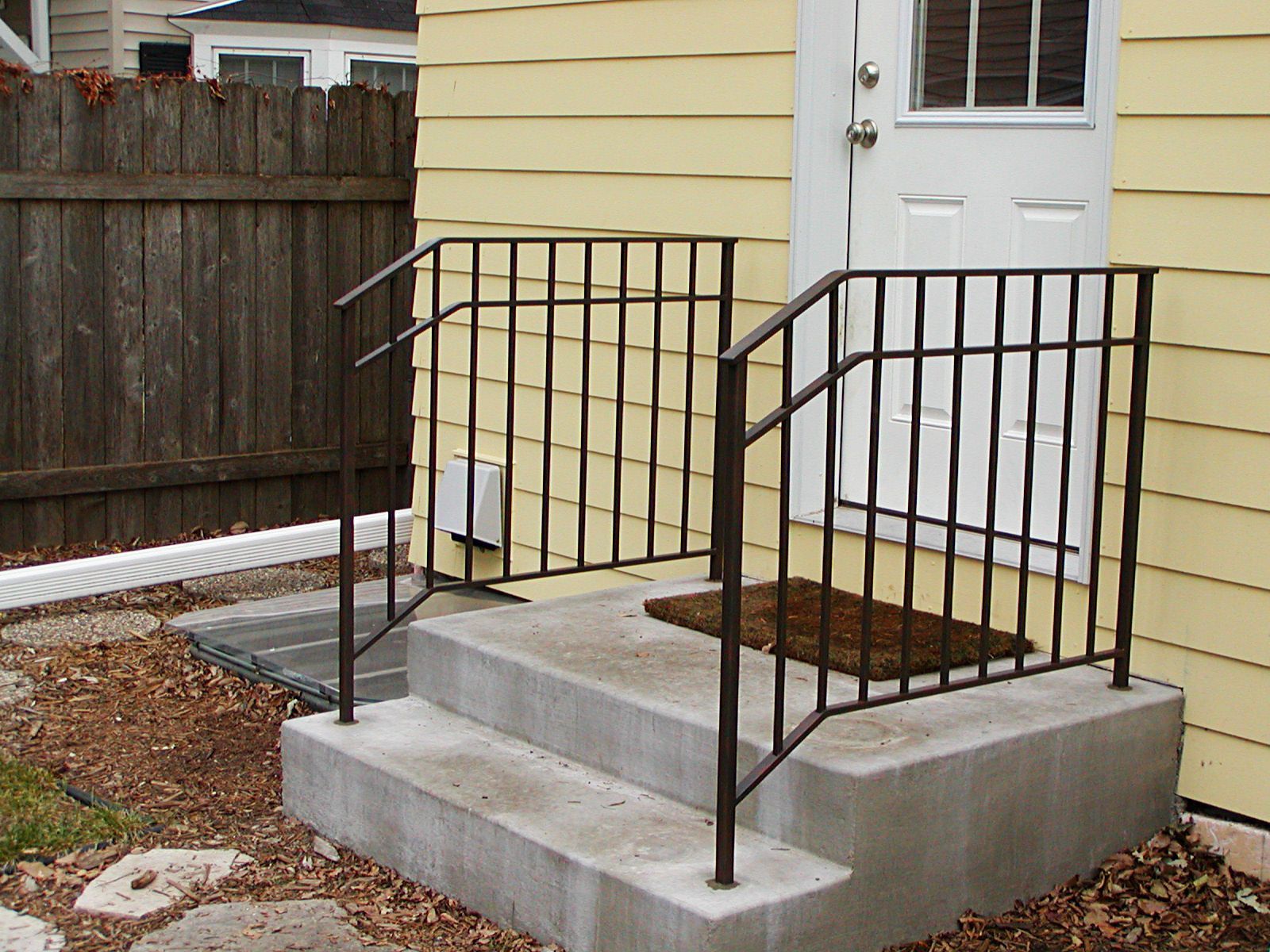 Concrete steps with metal handrail leading to a yellow-sided door; brown welcome mat.