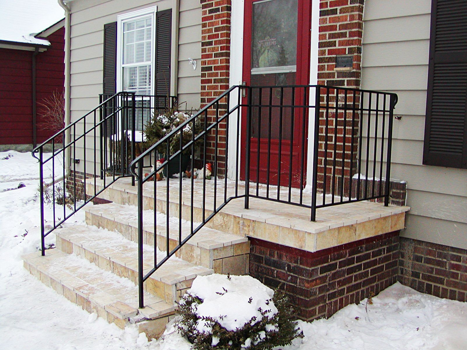 Black metal railings on brick and stone steps leading to a red door, snow on the ground.