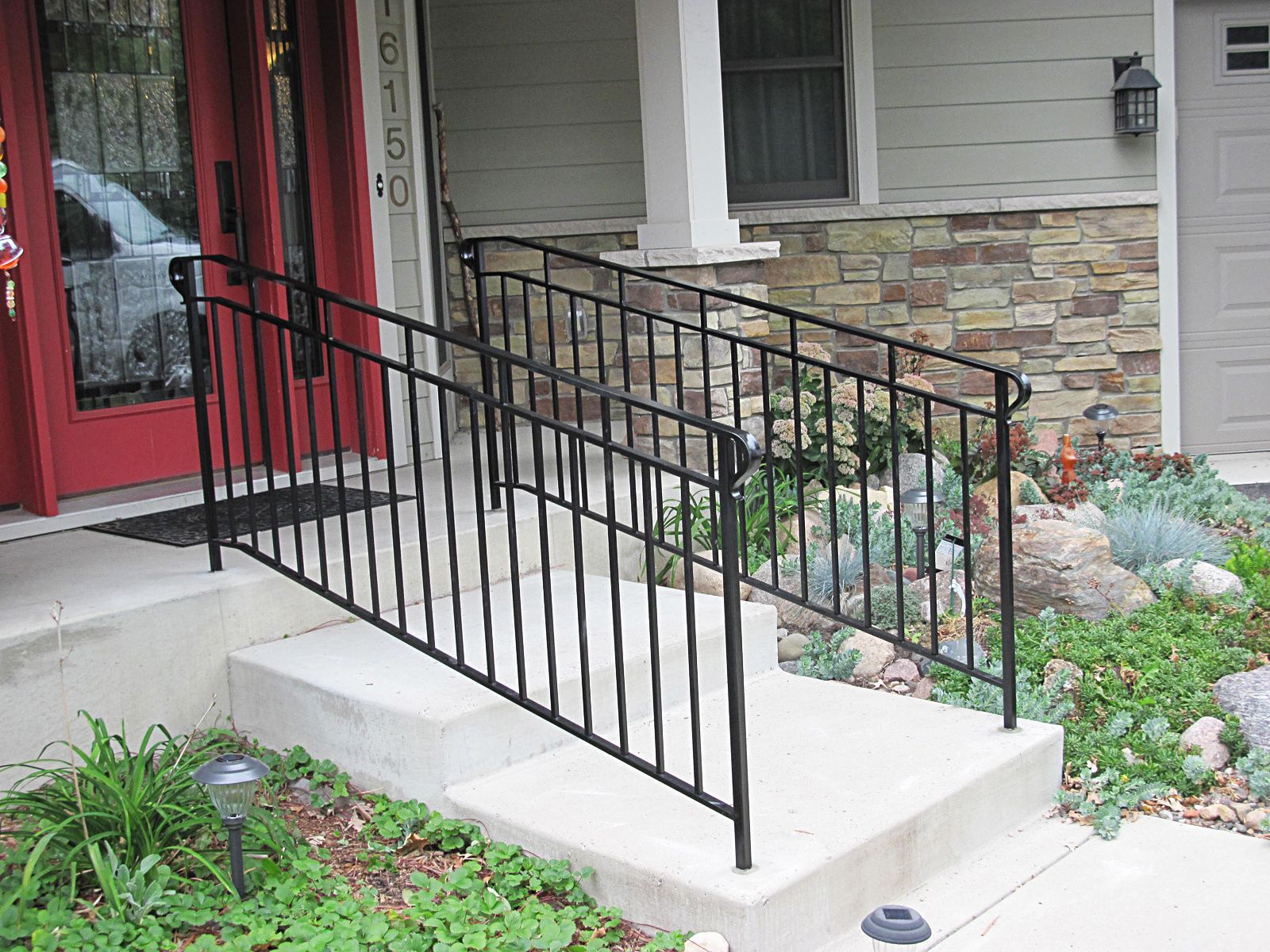 Black metal handrails on concrete steps leading to a house entrance. Green plants surround the steps.