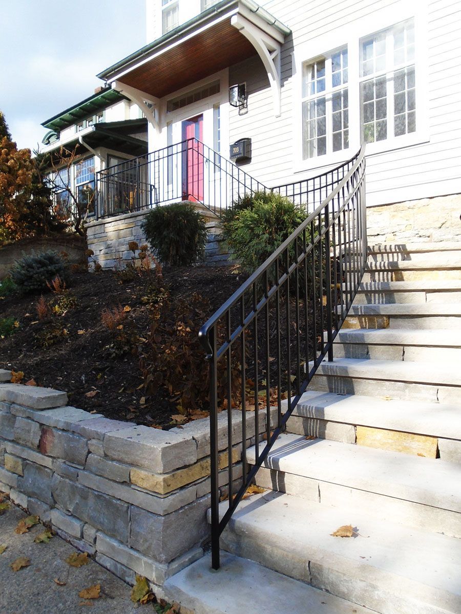 Exterior view of a house with stone steps and black railing leading to a red door. Landscaping and a covered porch are also visible.