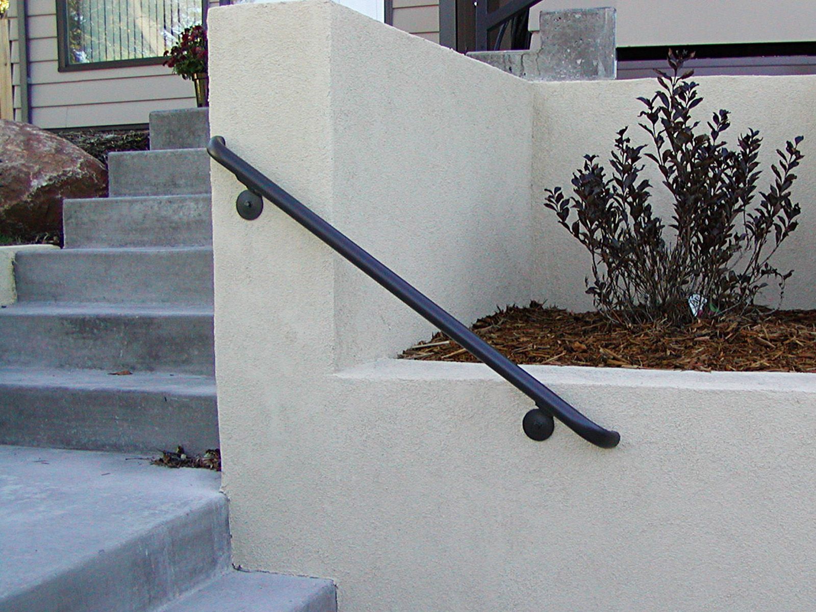 Black handrail on light stucco wall next to outdoor concrete stairs and a small bush.