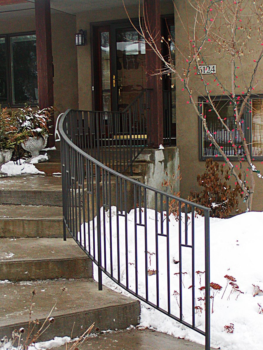 Black iron railing curving up concrete steps to a building entrance; snow on the ground.