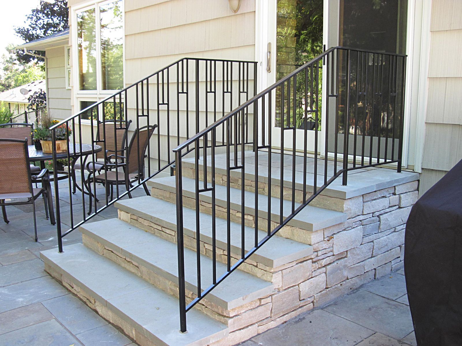 Black metal handrails on gray stone steps leading to a door, beside a patio.
