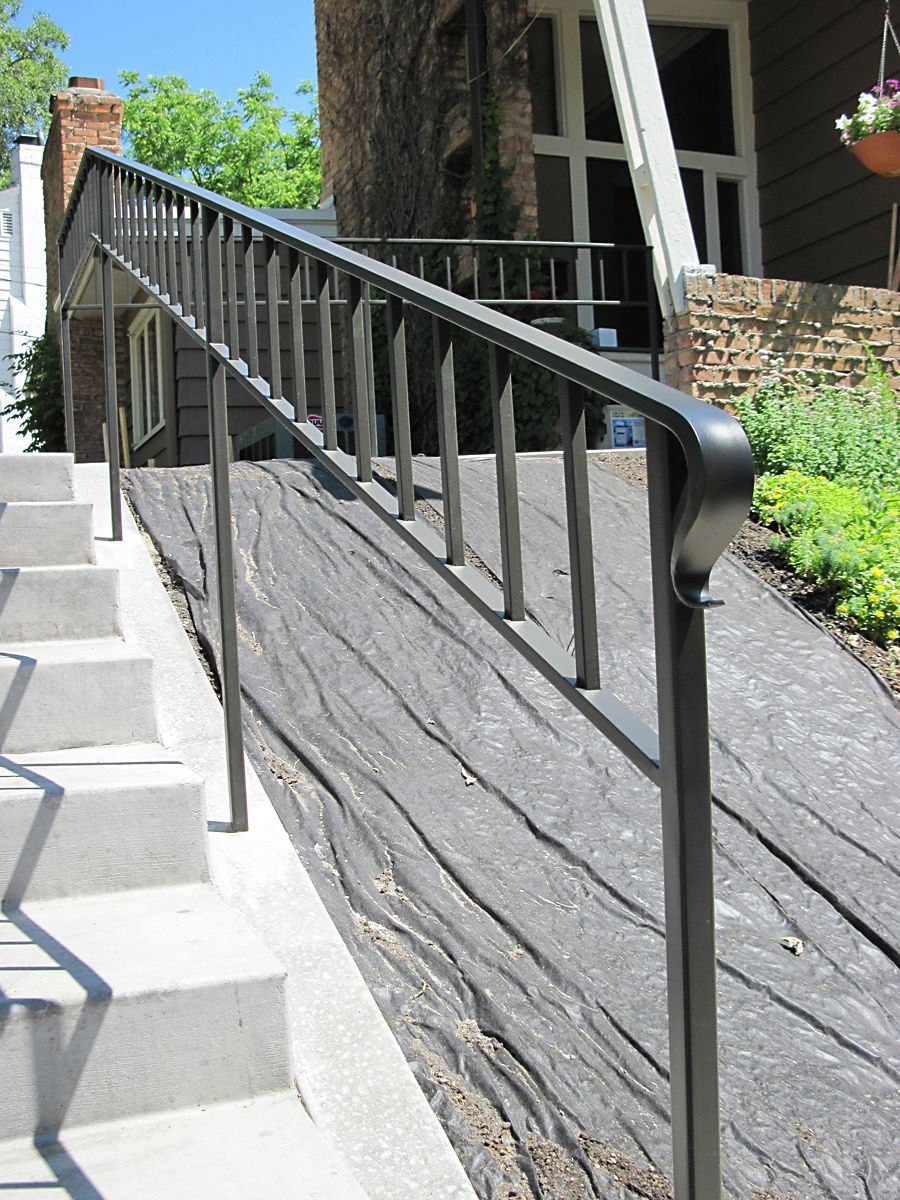 Black metal handrail on a ramp with textured wooden boards and concrete steps leading to a house.