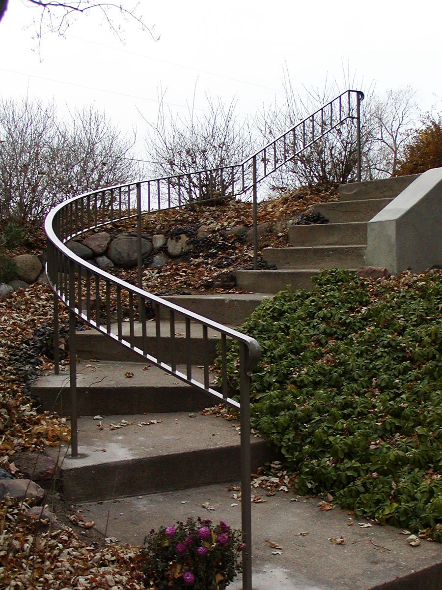 Concrete stairs with curved metal railing leading up a hillside, surrounded by greenery and fallen leaves.
